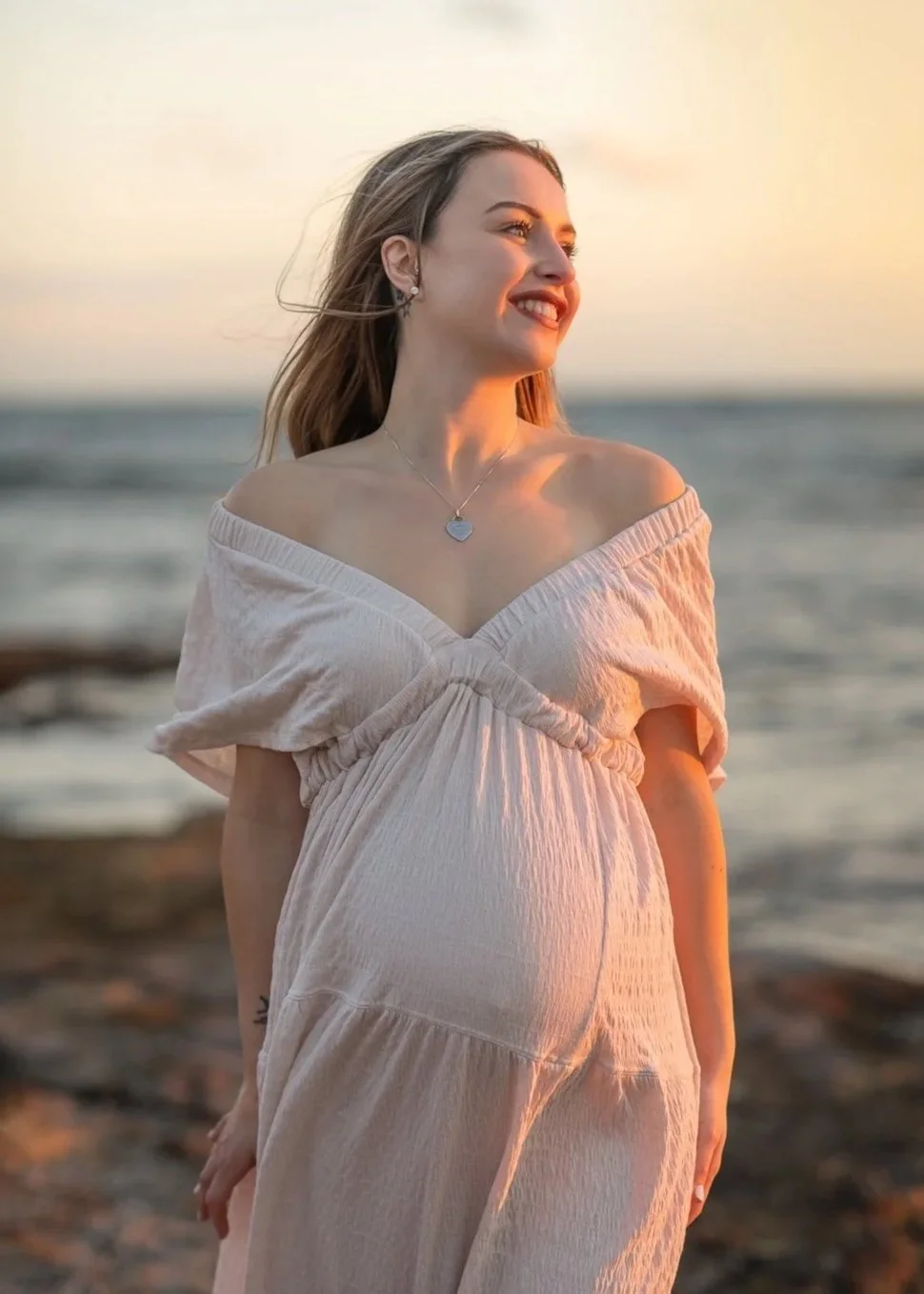 A young woman smiling on a beach during sunset, wearing a light-colored off-shoulder dress and a heart-shaped necklace.