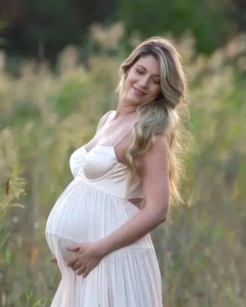 A pregnant woman with long, wavy blonde hair in a white dress standing in a natural outdoor setting, gently holding her belly and smiling with her eyes closed.