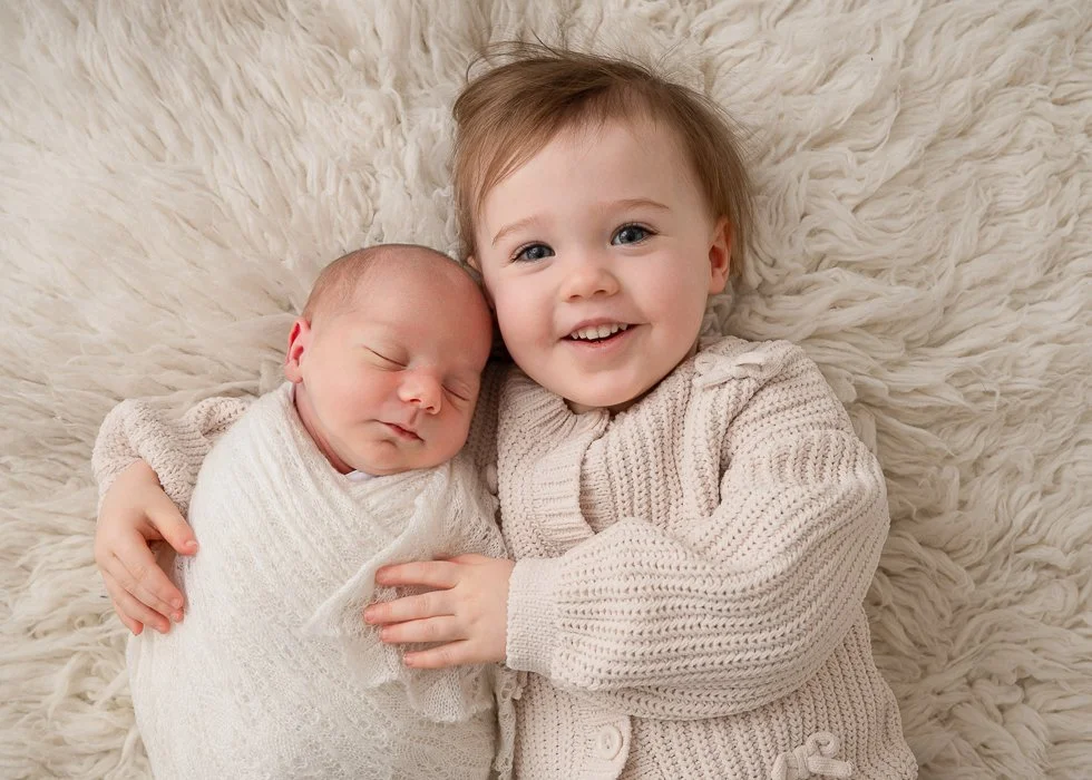 A young boy with light brown hair and blue eyes smiling while holding a sleeping newborn wrapped in a white blanket on a fluffy cream-colored rug.