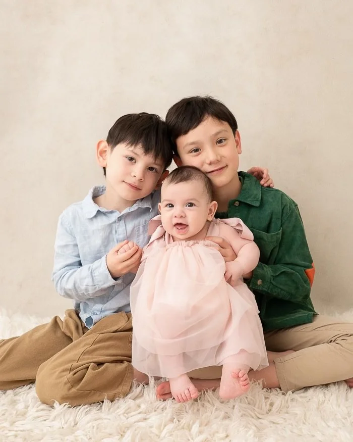 Studio image of two brothers looking at the camera holding their baby sister who wears a pink dress
