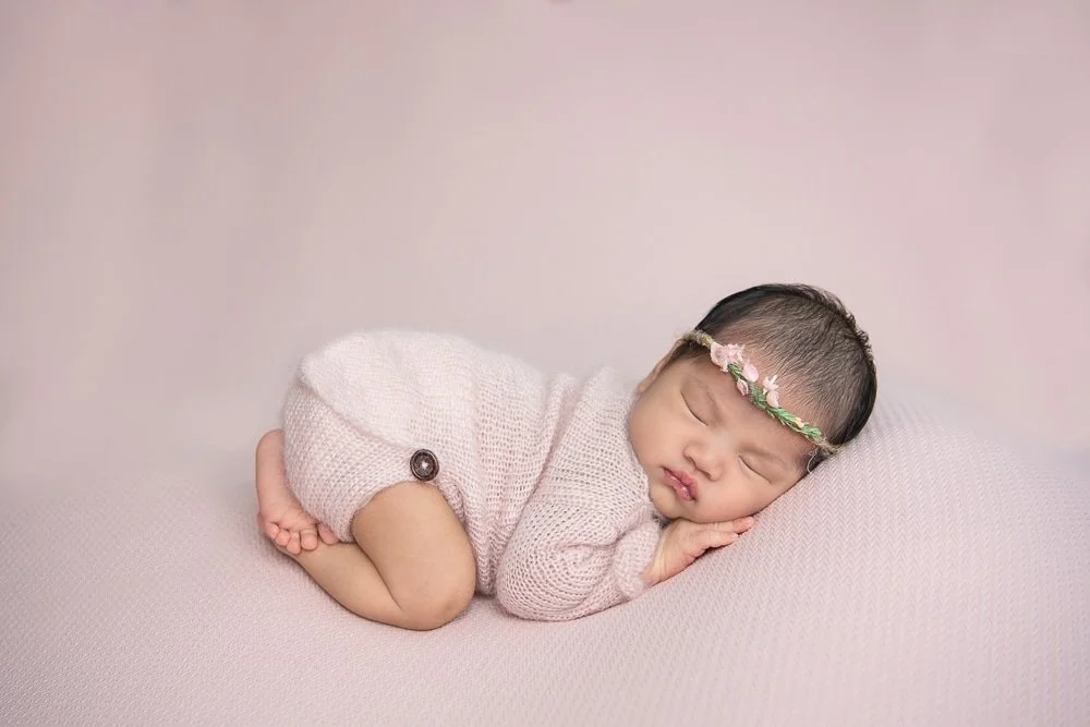 Newborn girl sleeping on pink blanket, wearing a knit outfit and floral headband.