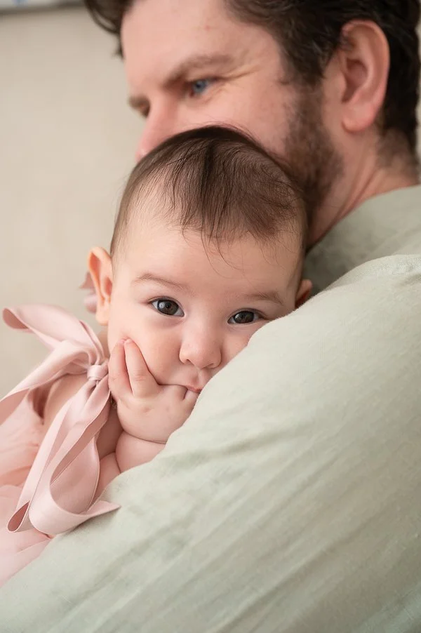 Melbourne baby photographer image of a baby sucking her fingers while being cuddled by her dad
