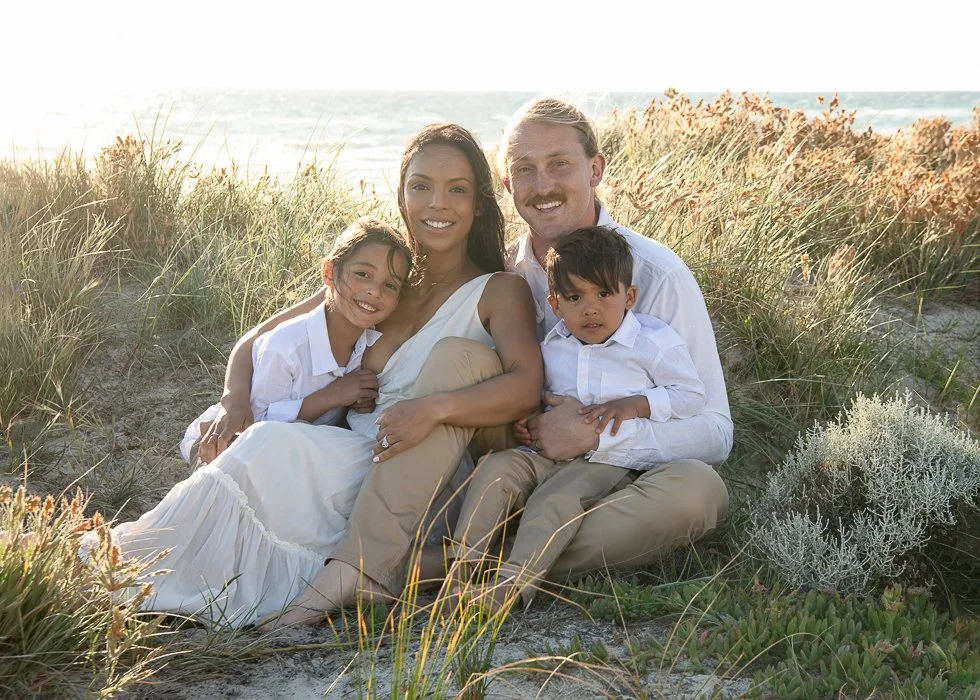 A family of four sitting on the beach surrounded by grass and plants during sunset.
