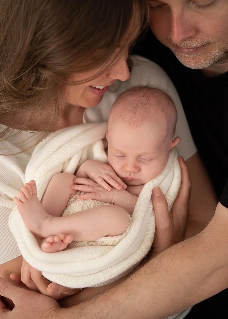 A smiling woman and a man looking at a sleeping baby wrapped in a cream blanket.