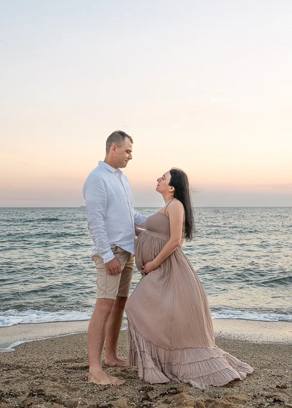 Melbourne maternity photography image of a woman in a flowing pink dress holding her belly, she is with her partner and the sun is setting so the sky is coloured pink.