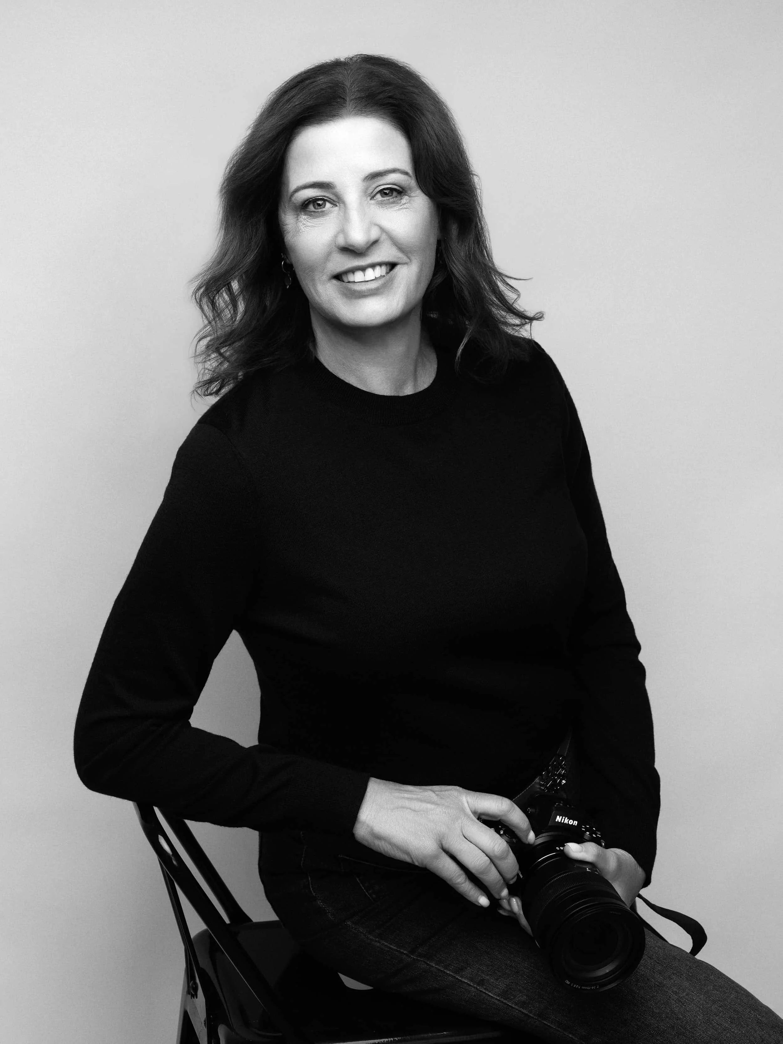 Image of Melbourne photographer Susan Bradfield monochrome, sitting on a chair smiling at the camera