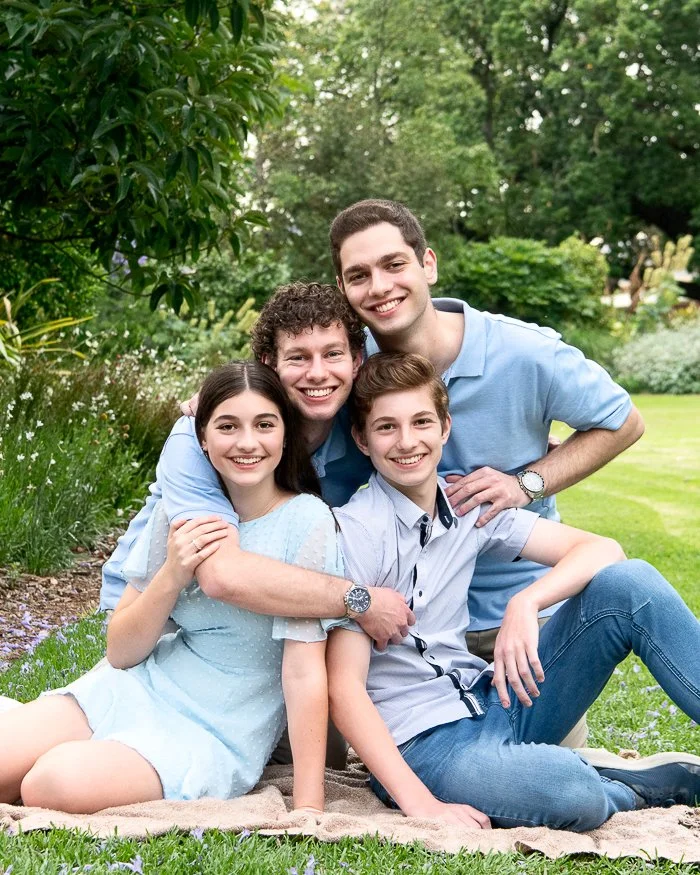 A family of five sitting on a blanket in a lush garden, smiling at the camera. The family includes two young children, a teenage boy, and two young adults, possibly their parents. The setting features green trees and plants.