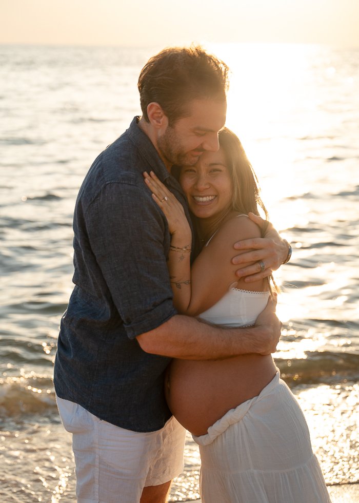 A couple hugging on the beach at sunset, with the woman being pregnant, both smiling happily.