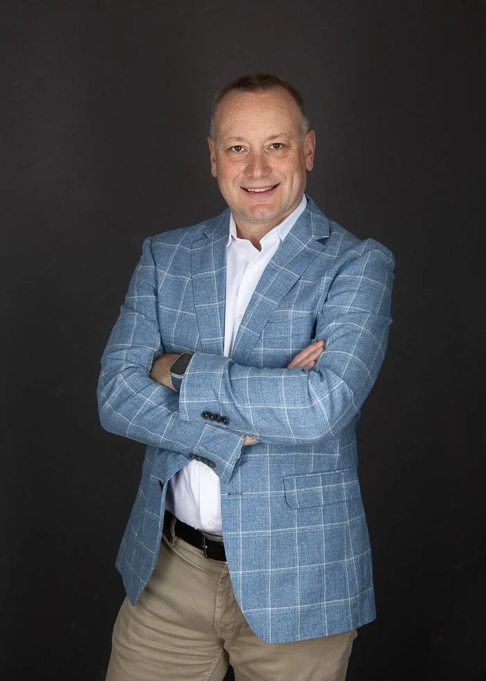 Susan Bradfield Photography headshot image of a man in a blue check blazer standing with his arms folded