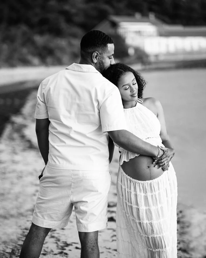 Monochrome of a pregnant woman at the beach leaning into her partner who is holding her and her tummy