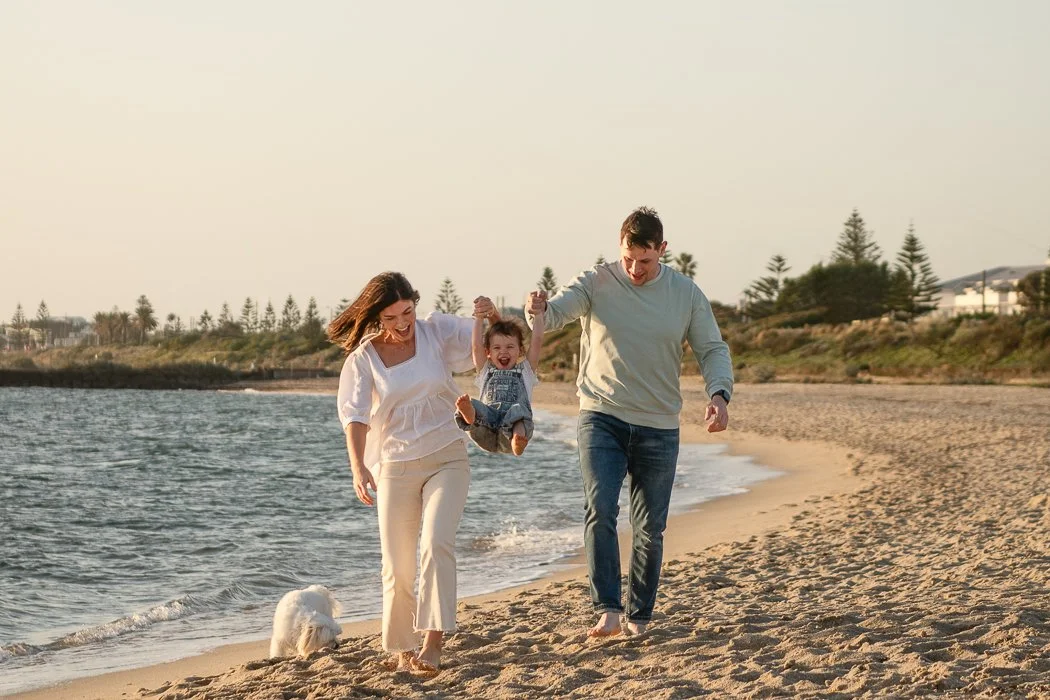 Melbourne bayside family photographer image of a family swinging their your child between them . There is a white dog and the child is very happy at the beach