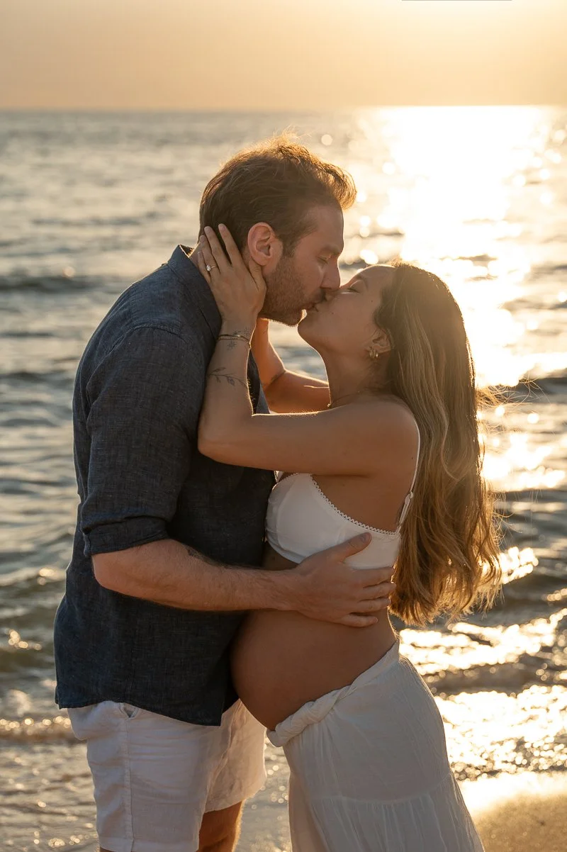Photo of a pregnant woman and her partner kissing at sunset at the beach
