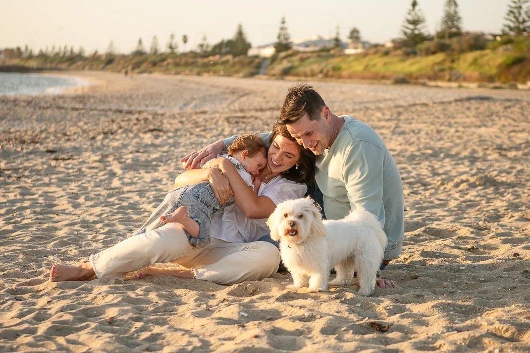 Melbourne family photographer photo of parents sitting at the beach hugging their little daughter. A white small dog looks at the camera