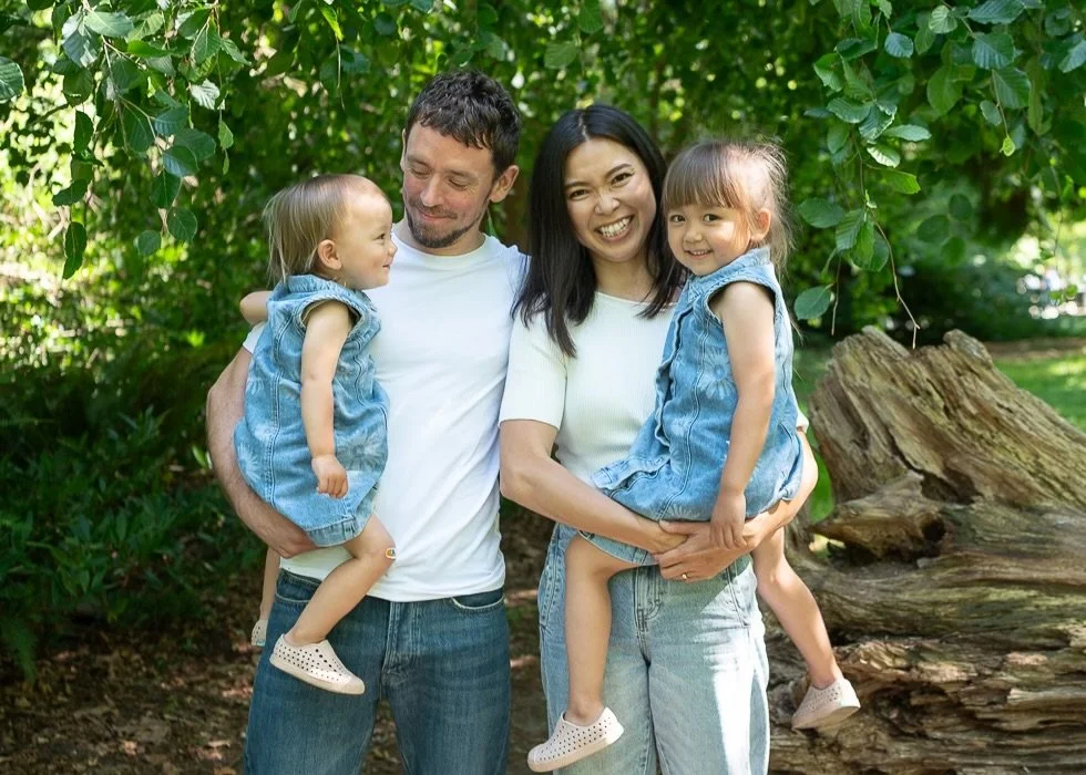 Happy parents with their two young daughters wearing denim dresses picture outdoors under a tree