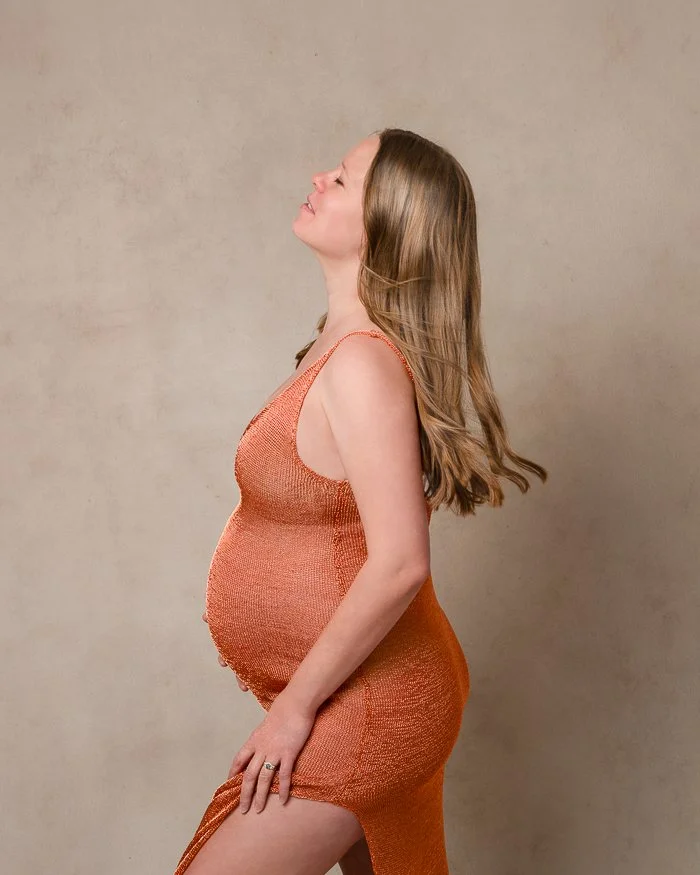 Pregnant woman with long hair wearing an orange dress, standing in profile against a neutral background.