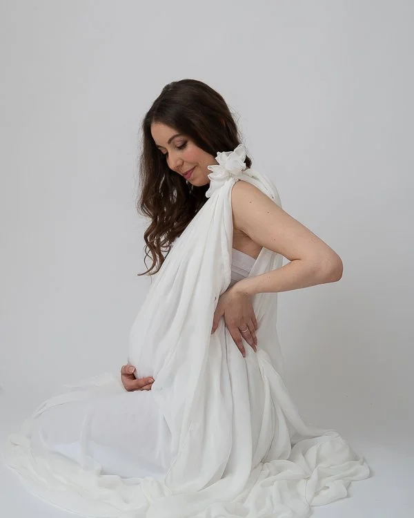 Melbourne photographer Studio photo of pregnant woman side on wearing a white outfit and kneeling with a white backdrop 