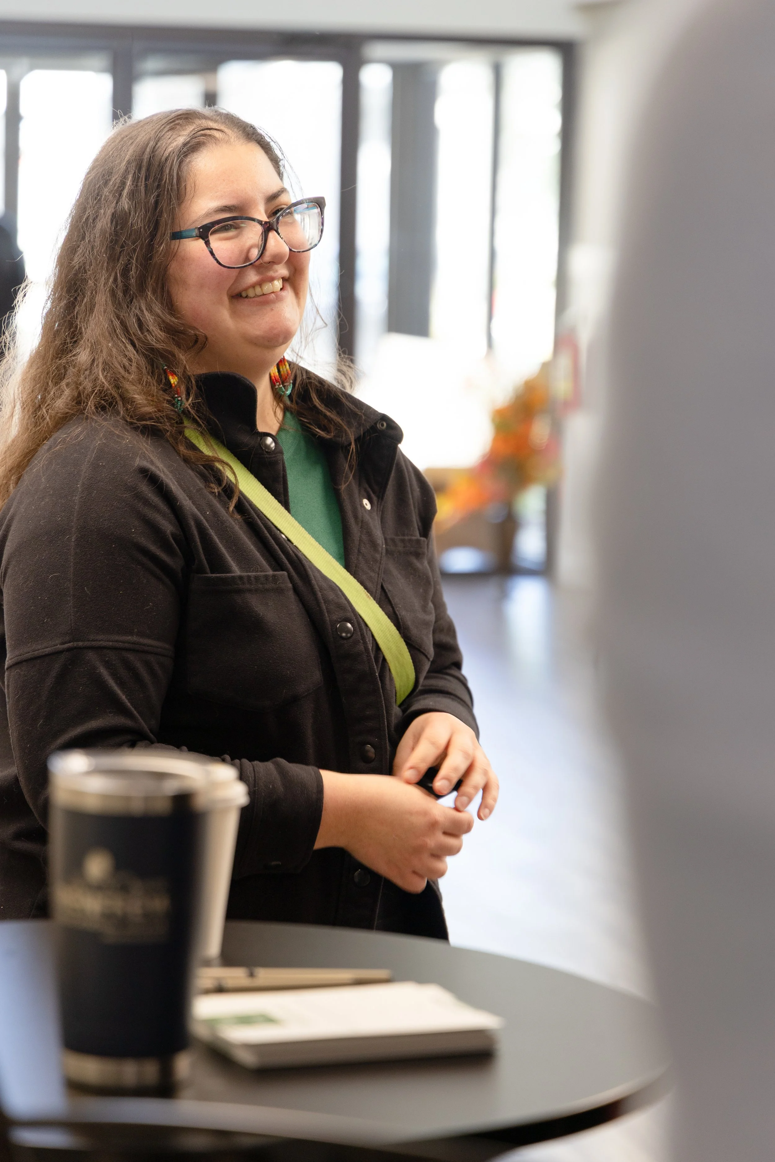 A woman with long brown hair, glasses, and colorful earrings smiling while standing indoors near a window with blurred background. She is wearing a black jacket over a green shirt, with a green strap across her chest.
