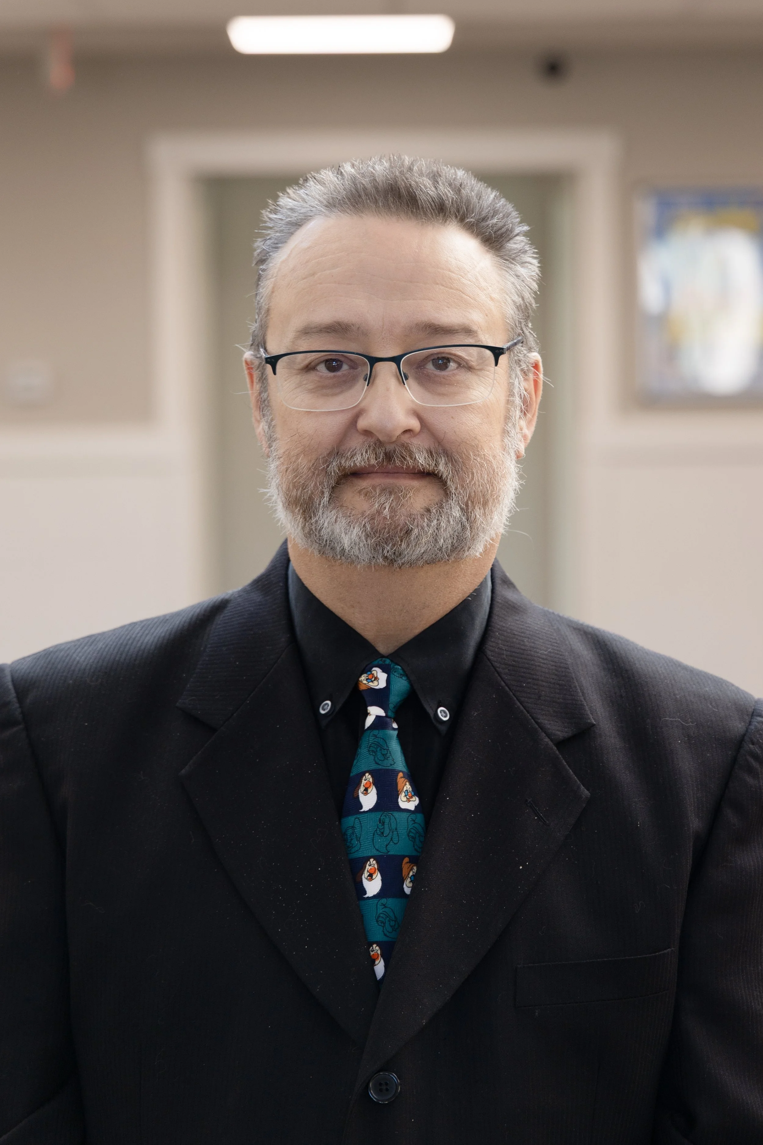 A man with glasses, gray beard, and gray hair wearing a black suit, black shirt, and a tie with penguins, standing indoors in front of a blurred background.