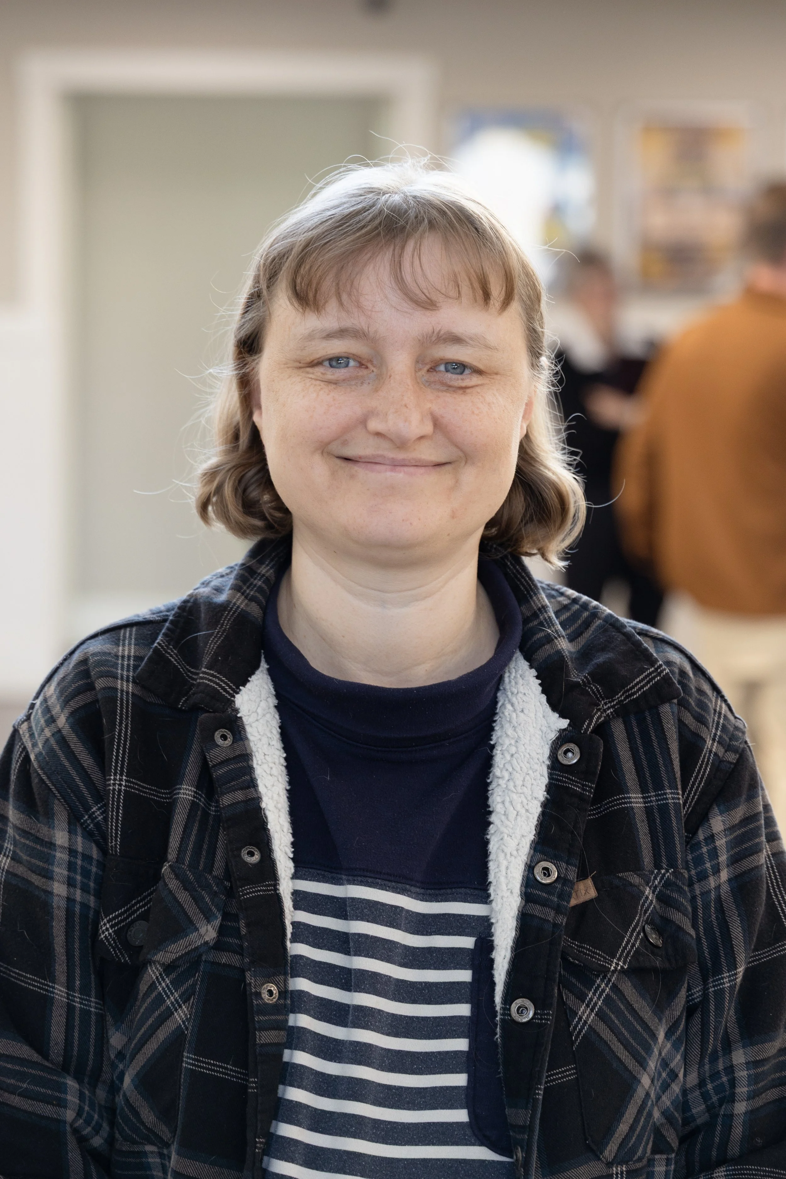 A person with light skin, blue eyes, and short light brown hair, smiling at the camera. They are wearing a dark striped shirt under a plaid jacket with fleece lining, in an indoor setting with blurred people and windows in the background.