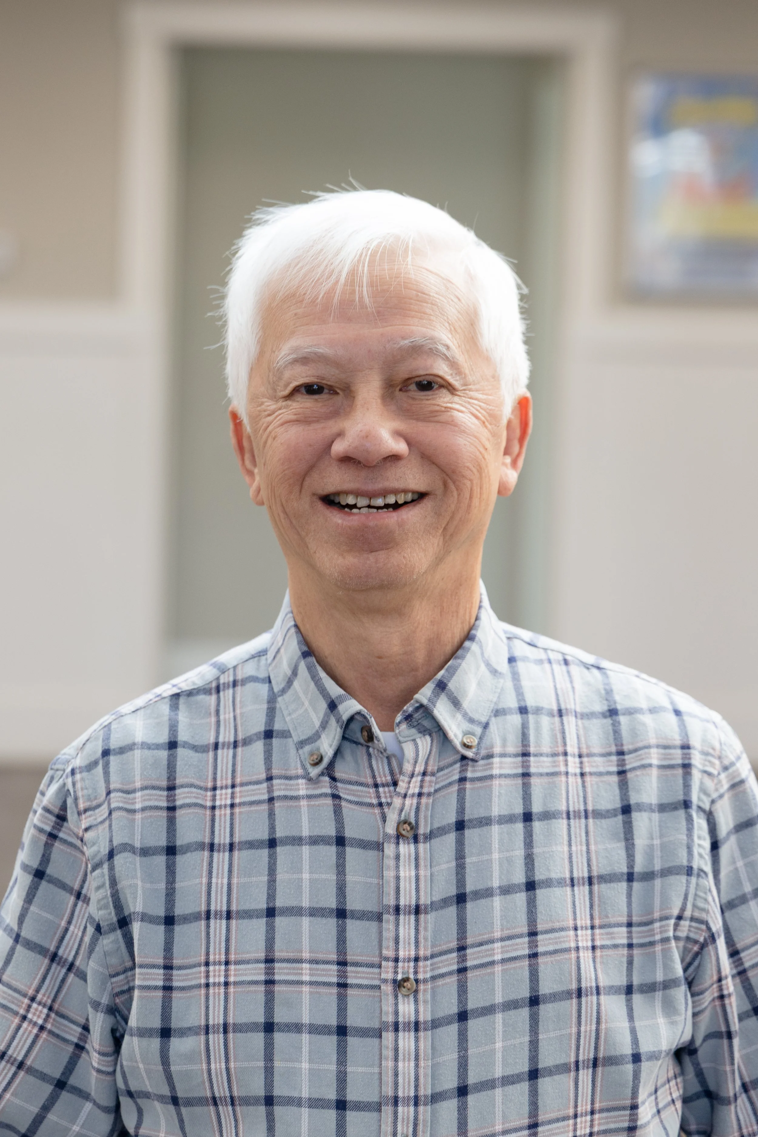An elderly man with white hair smiling, wearing a blue and white plaid shirt, standing indoors with blurred background.