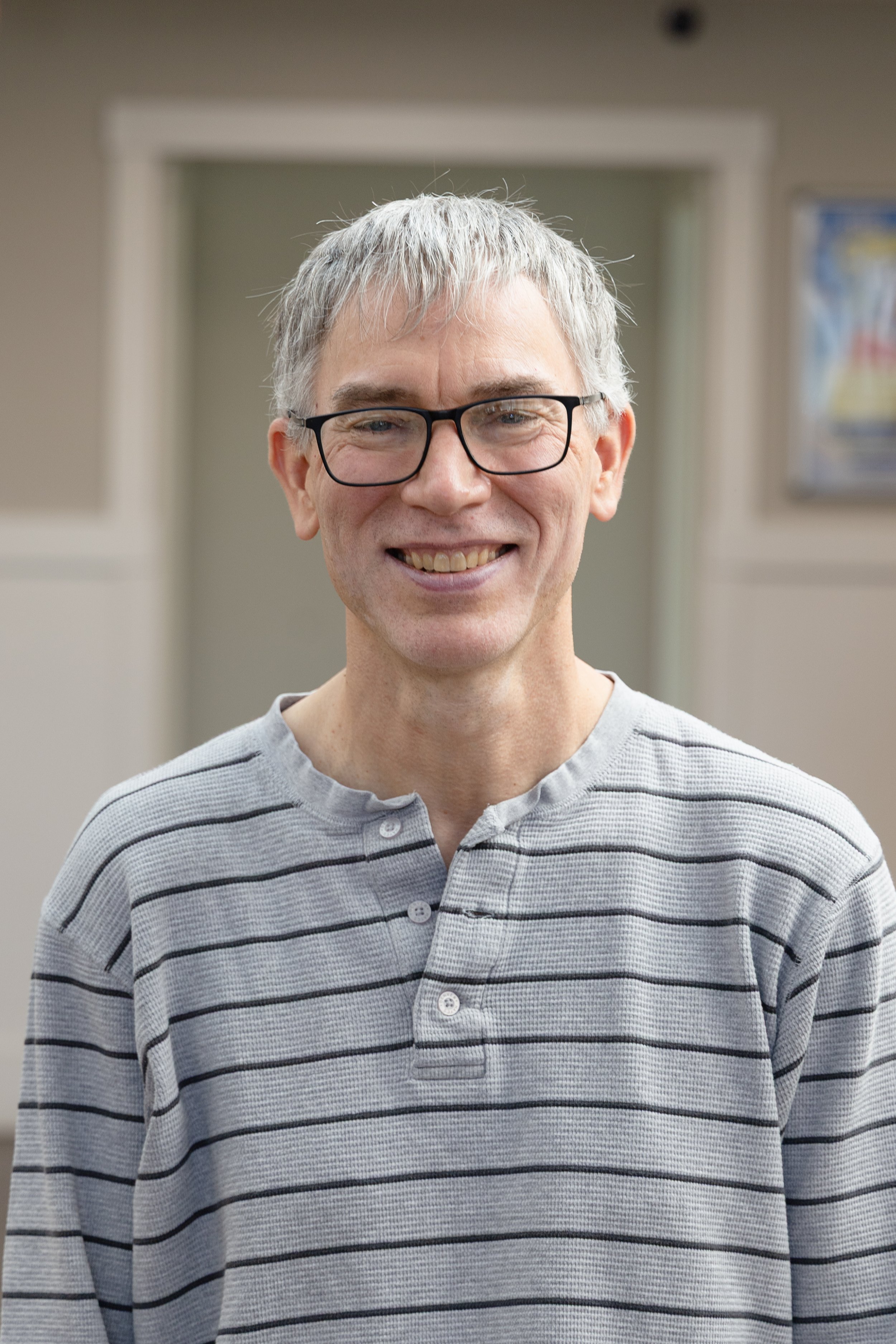 A man with gray hair and glasses wearing a gray and black striped shirt, smiling in an indoor setting.