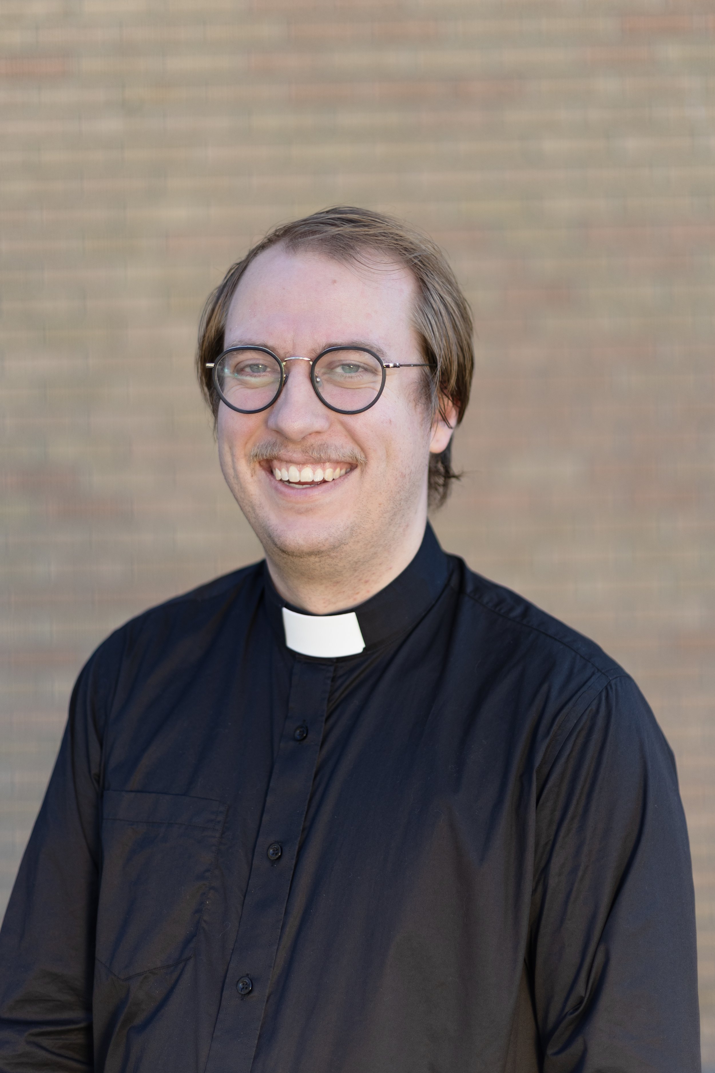 Portrait of a young man with glasses, smiling, wearing a black clerical shirt with a white collar, standing outdoors against a blurred brick wall background.