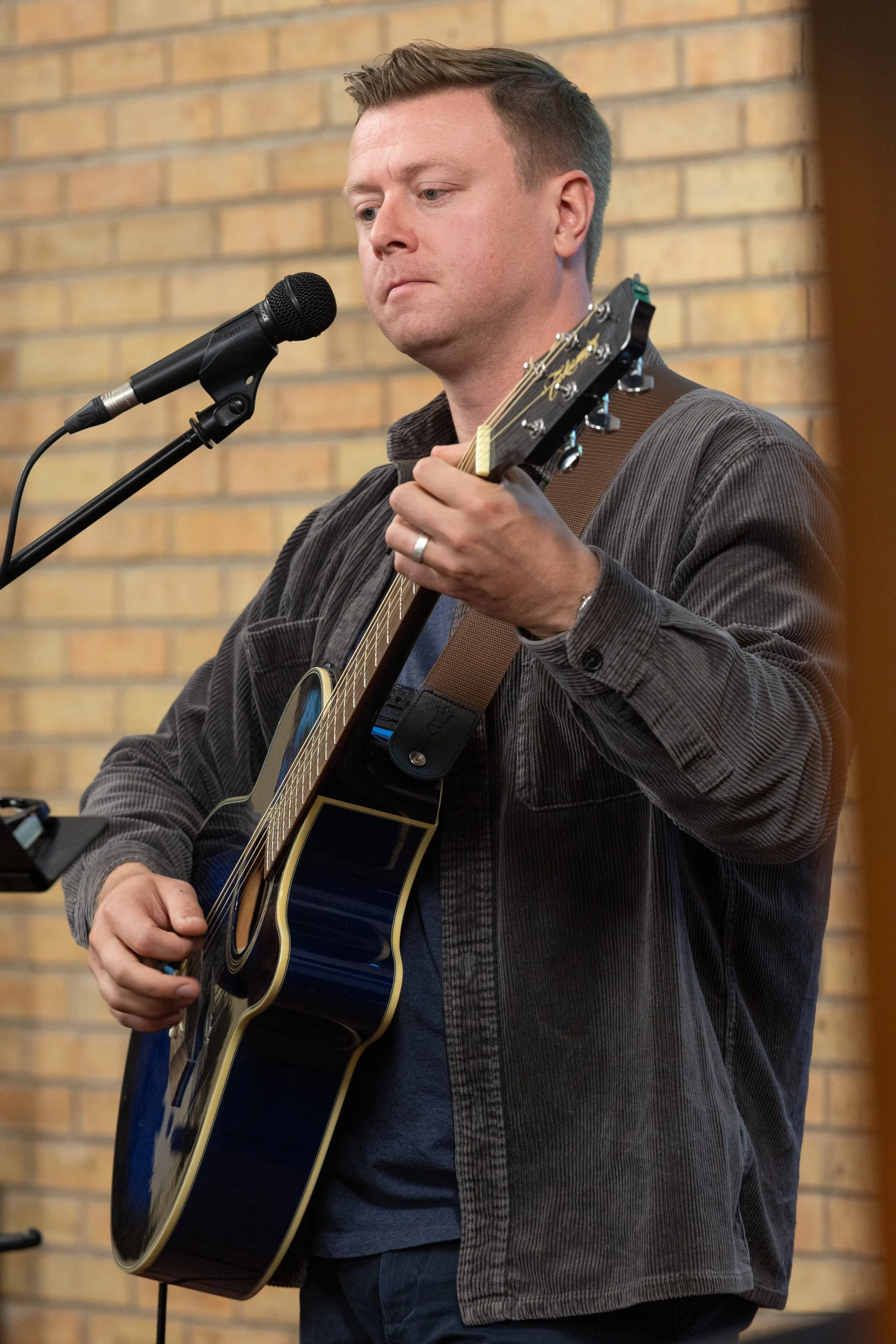 A man playing an acoustic guitar into a microphone against a brick wall background.