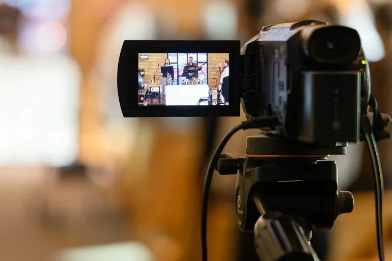 Video camera on a tripod recording a live musical performance, visible on its flip-out screen