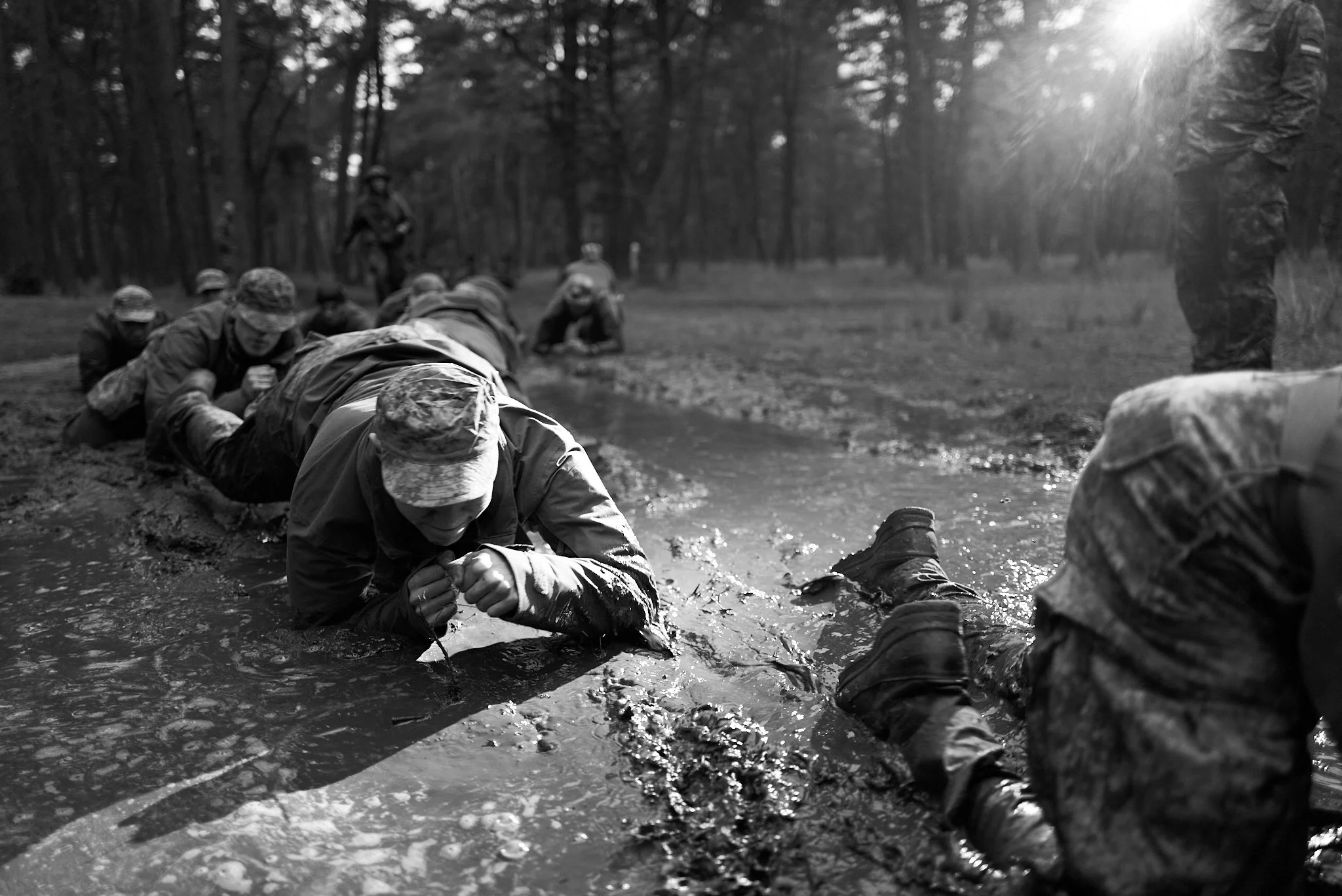 Mariniers tijgeren door een modderige waterplas tijdens een intensieve training in een bosrijk gebied, vastgelegd in zwart-wit, met nadruk op fysieke inspanning, doorzettingsvermogen en samenwerking.