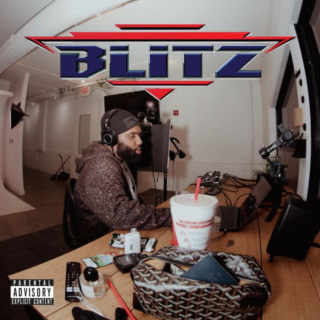 Man wearing headphones and a beanie working at a desk with audio equipment, a large cup, and personal items, in a studio or office setting, with a 'BLITZ' logo overhead.