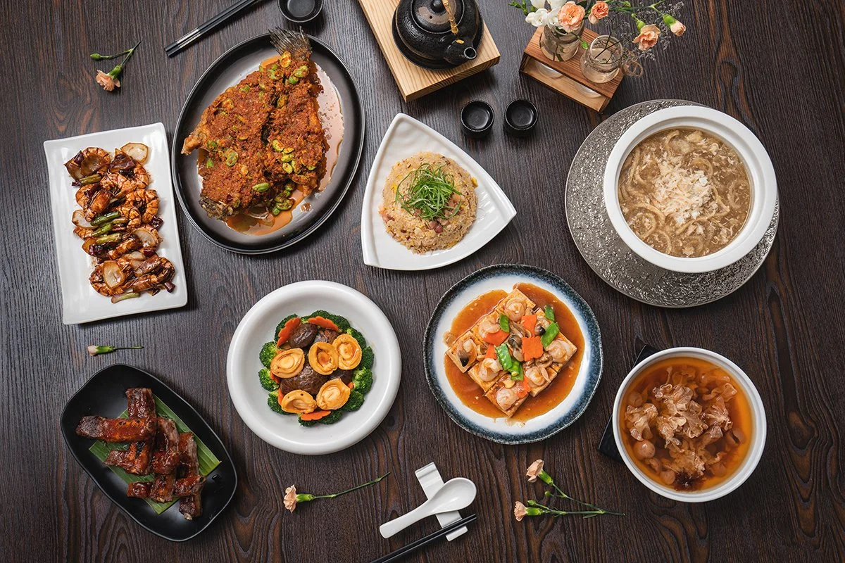 A variety of Chinese dishes on a wooden table, including a whole spiced fish, stir-fried eggplant, fried rice with scallions, soup, braised ribs, tofu with vegetables, and a floral teapot arrangement.