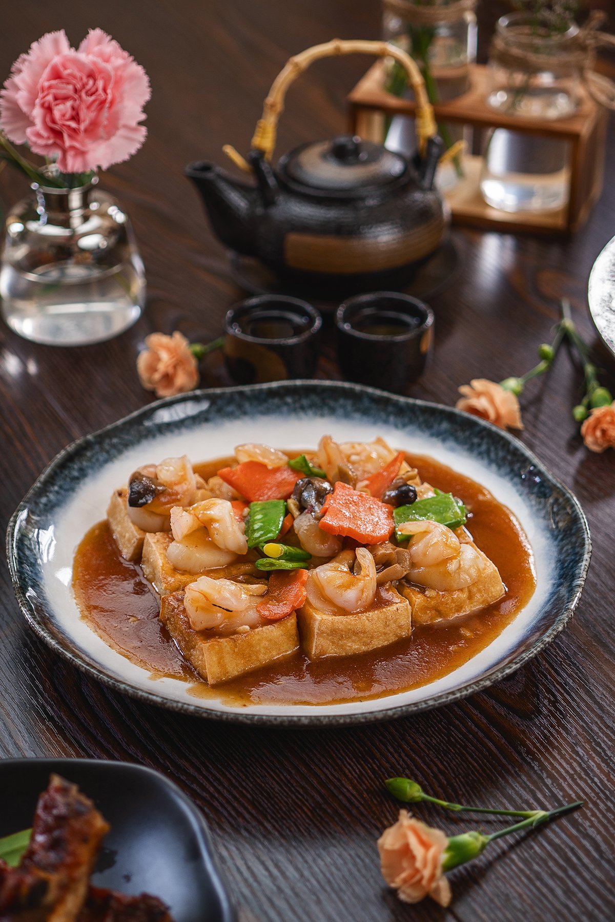 Plate of tofu with shrimp and vegetables in sauce, surrounded by pink carnations, a black teapot, and teacups on a wooden table.