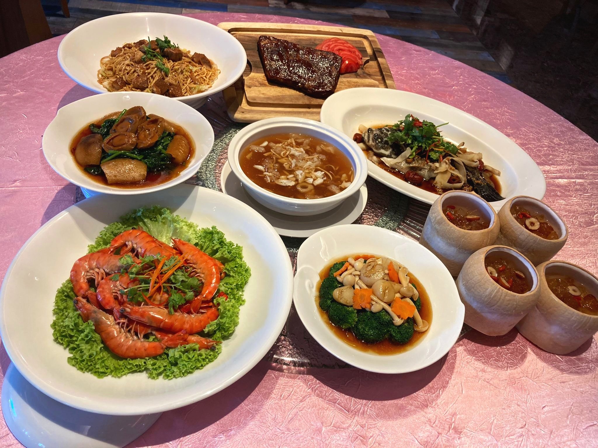 A variety of Chinese dishes on a table, including noodles with beef, braised vegetables, prawns on lettuce, soup, steamed fish with mushrooms, and herbal tea in cups.