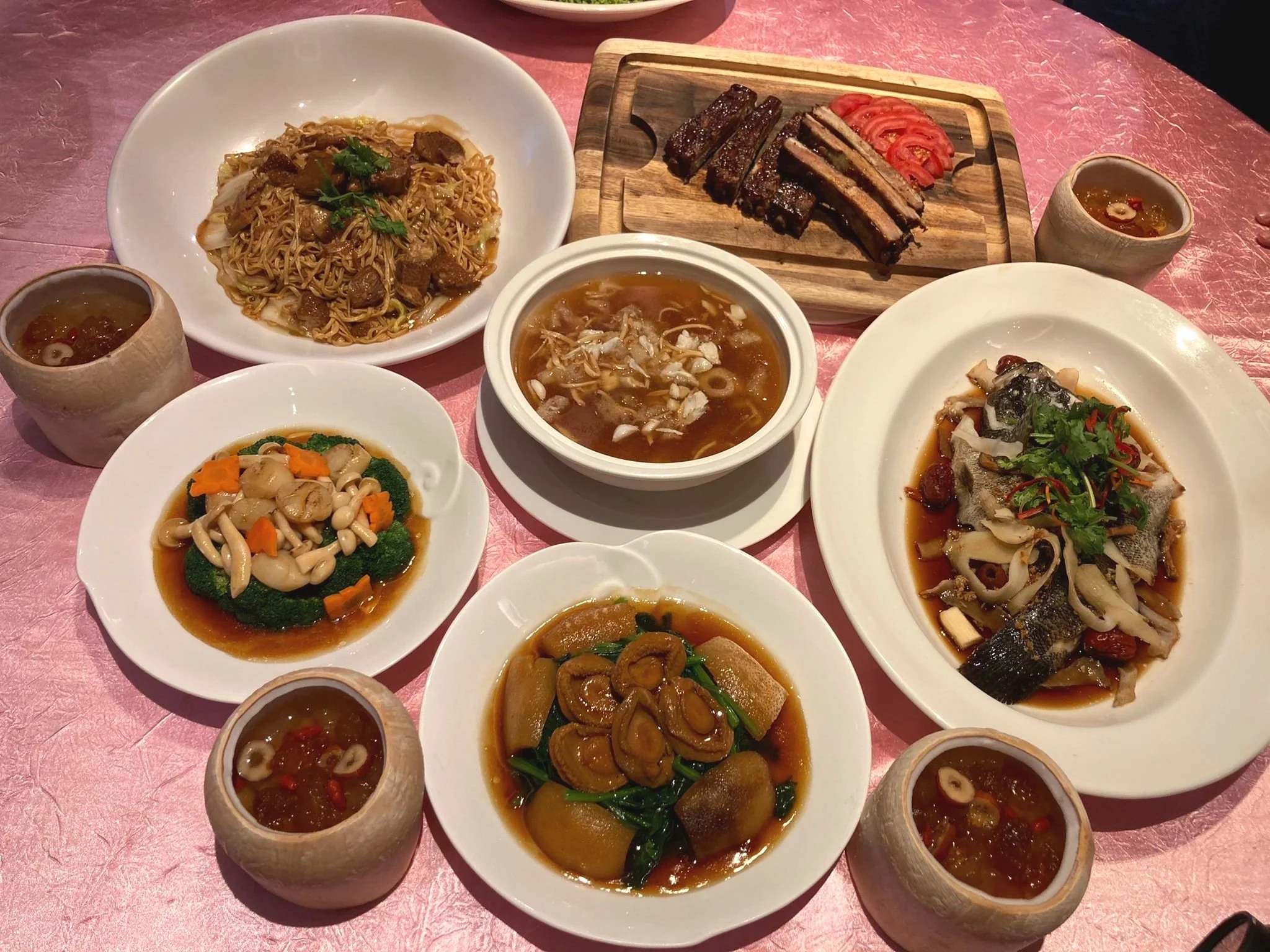 A variety of Asian dishes including noodles with meat, a clear soup, a dish with mushrooms and broccoli, ribs with sliced tomatoes, steamed fish with herbs, and other side dishes served with sauces in small cups, all on a pink tablecloth.