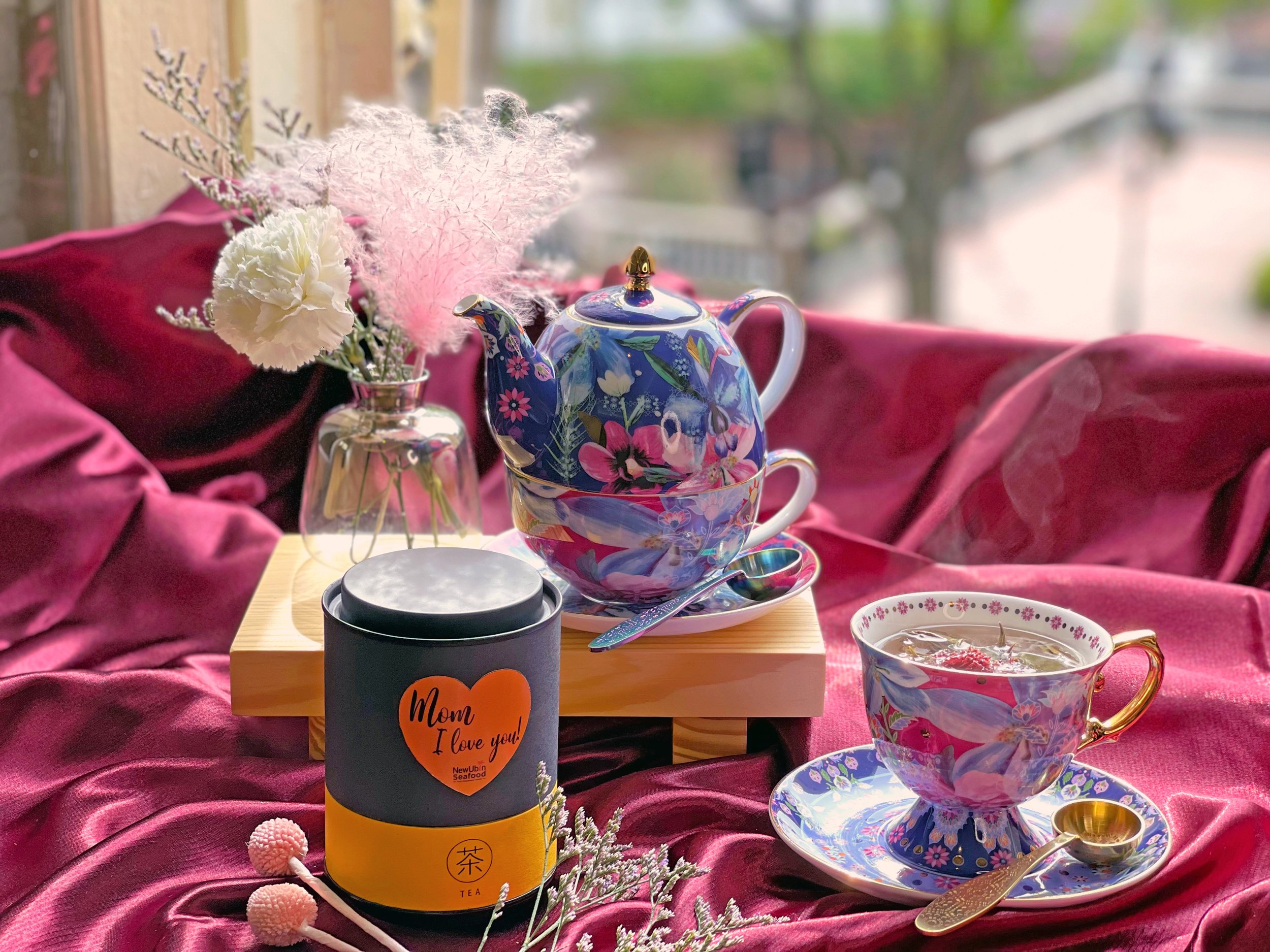 Floral teapot and teacup set with "Mom, I love you!" tea tin on a burgundy silk cloth, next to a vase with white and pink flowers.