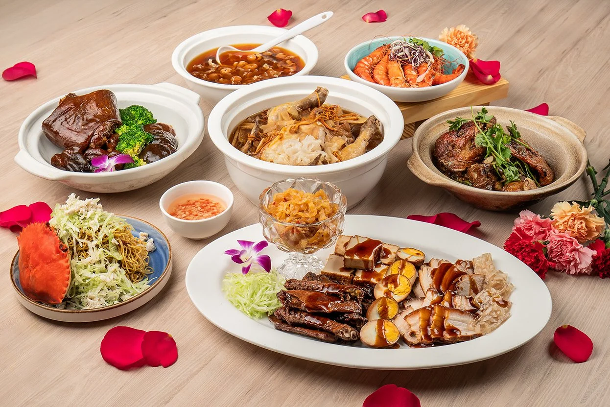 Assorted Chinese dishes on a table, including braised meat, steamed prawns, stir-fried noodles with crab, herbal soup with chicken, and tofu with tea eggs. Garnished with vegetables and flowers.