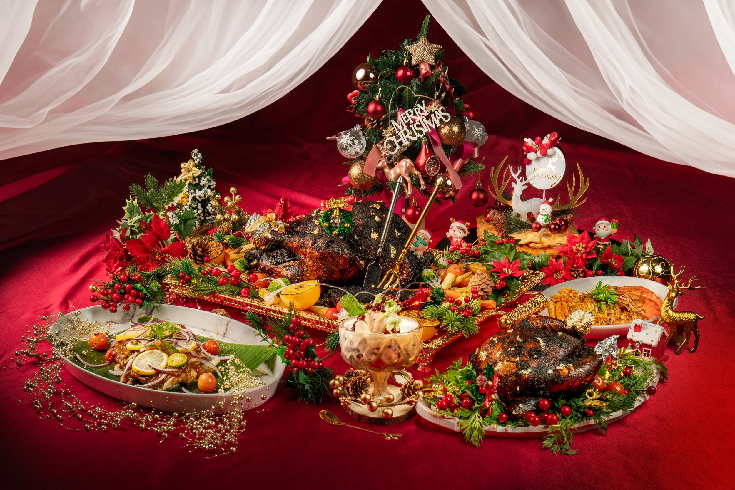 A festive Christmas dinner table featuring roasted meats, pasta, and desserts. The table is decorated with holiday ornaments, candles, and a miniature Christmas tree. Red and green decorations, including poinsettias and holly, enhance the festive atmosphere. A 'Merry Christmas' sign and seasonal figurines are also present.