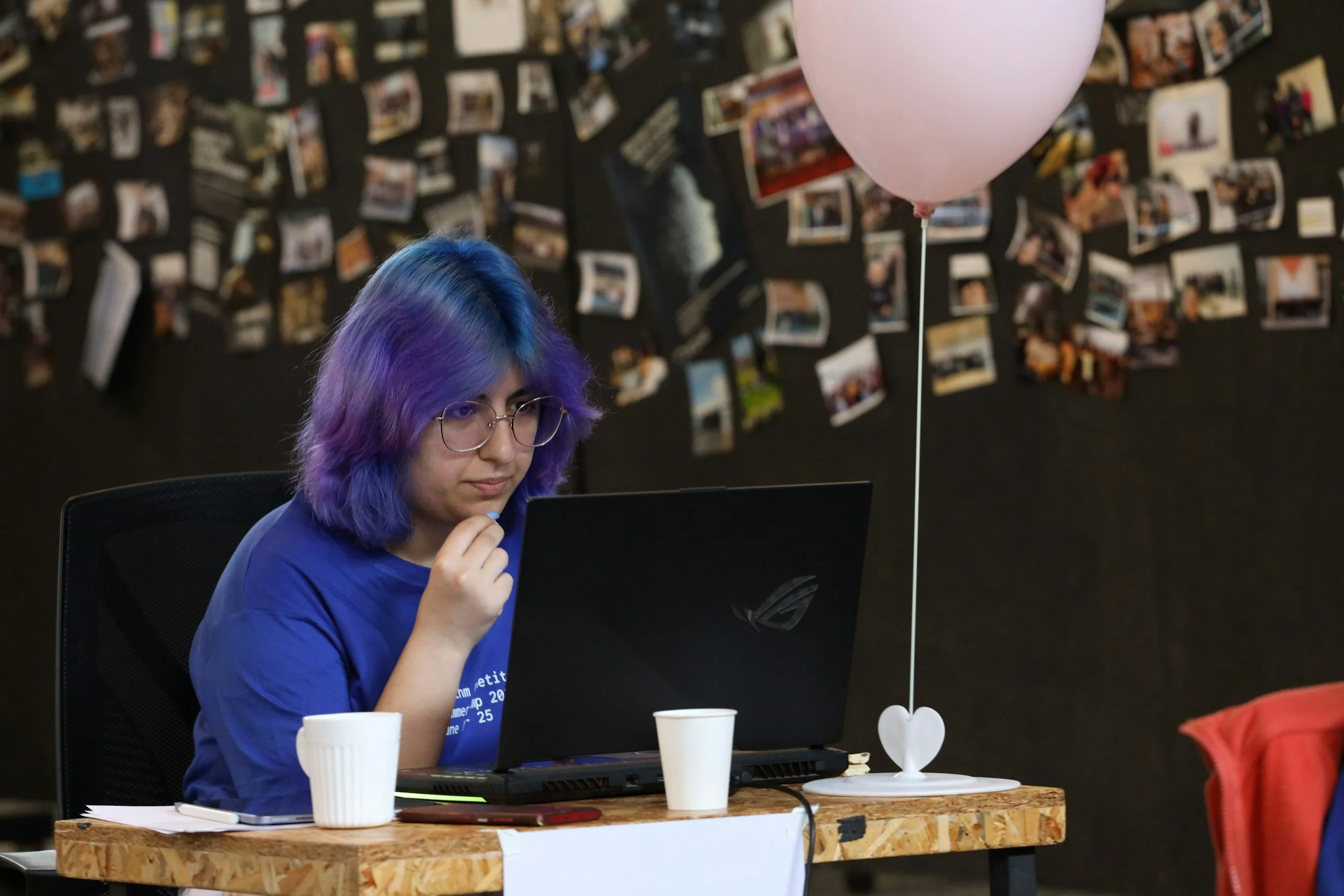 A young woman with purple and blue hair, wearing glasses and a blue shirt, working on a black laptop at a wooden table decorated with a pink balloon and a white cup. In the background, there is a black wall with numerous photos pinned to it.