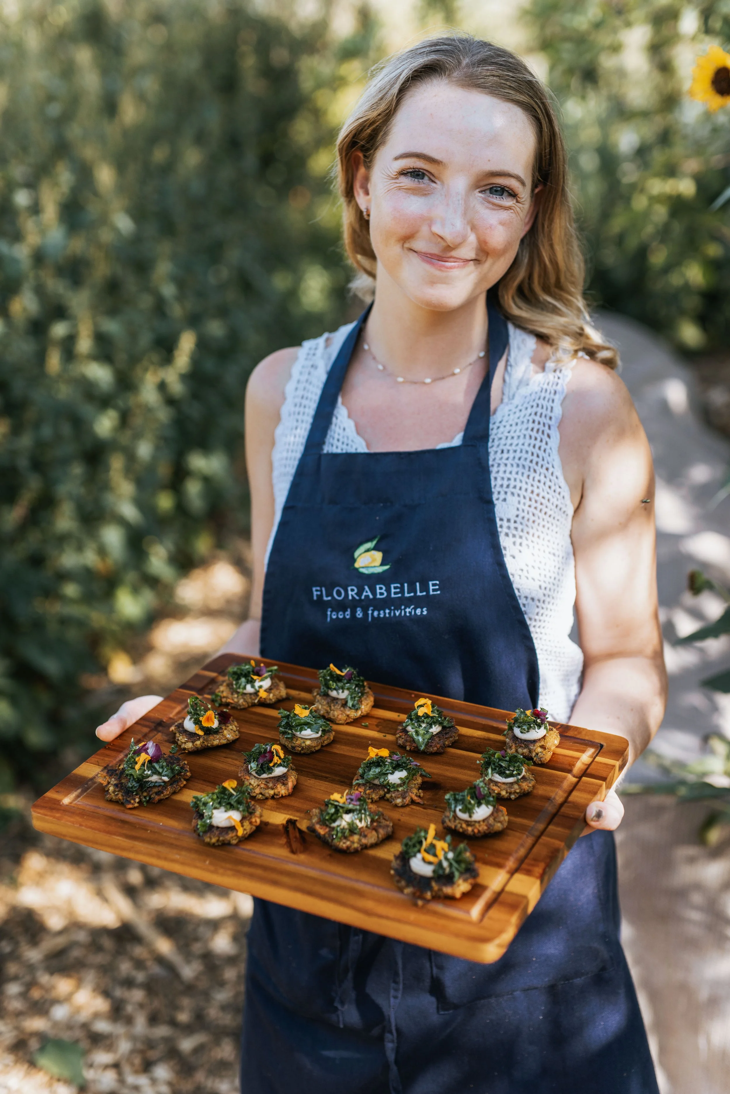 Chef Liza smiling and holding a wooden tray with small decorated appetizers outdoors. She is wearing a navy apron with the logo 'FLORABELLE food & festivities' and a white top, with trees in the background.