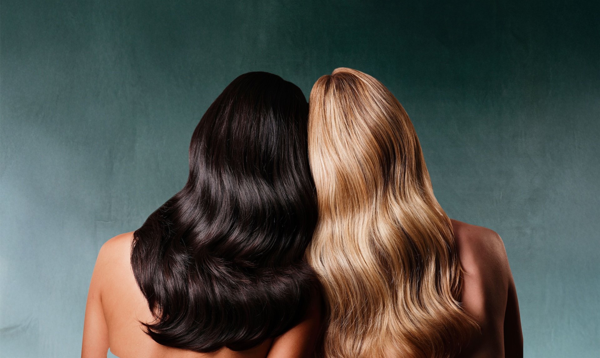 Two women with long, wavy hair, one with black hair and one with blonde hair, facing away from the camera and leaning their heads together.