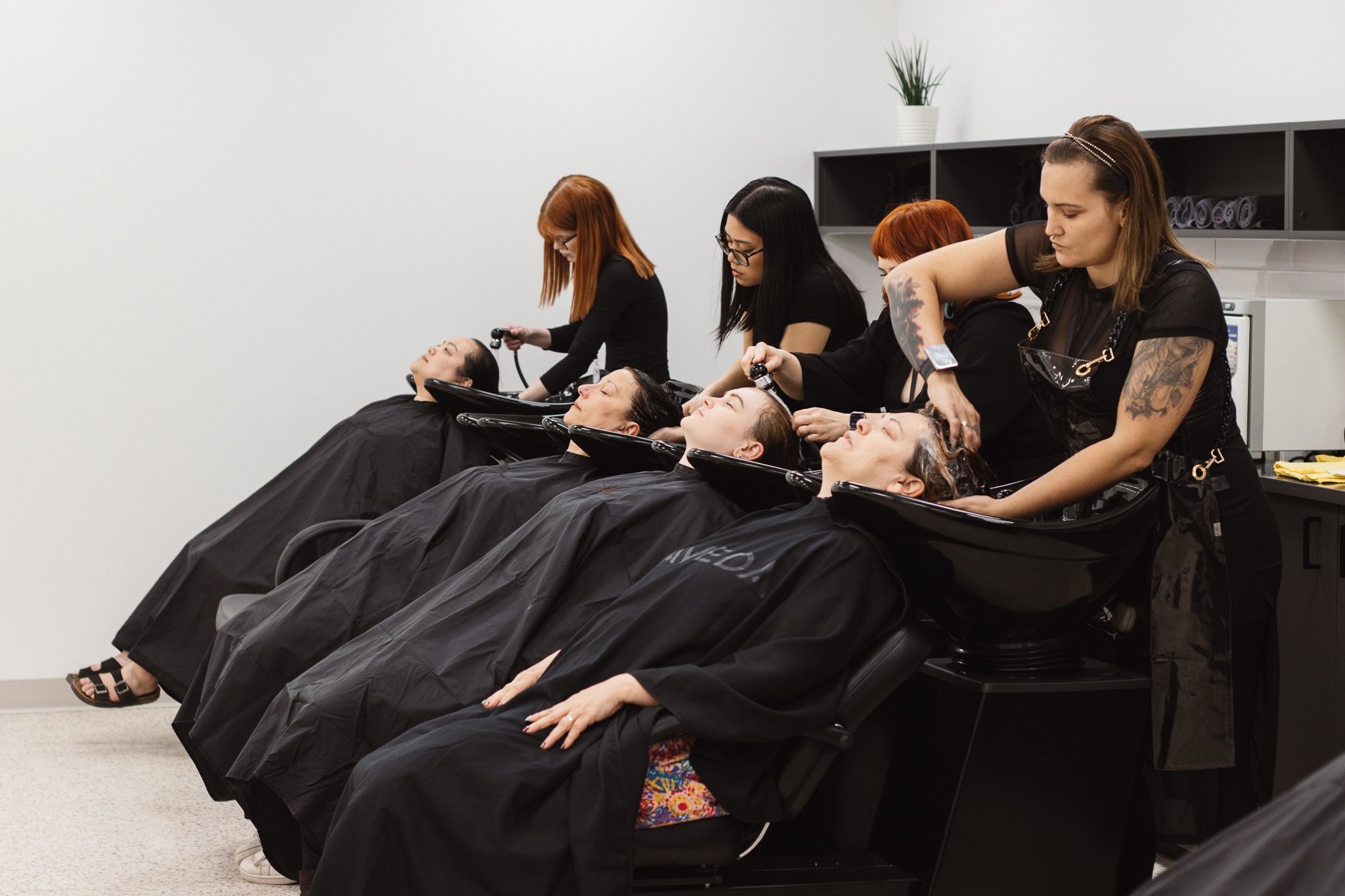 Four students washing their guests’ hair at salon sinks during a hands-on hairstyling training session.