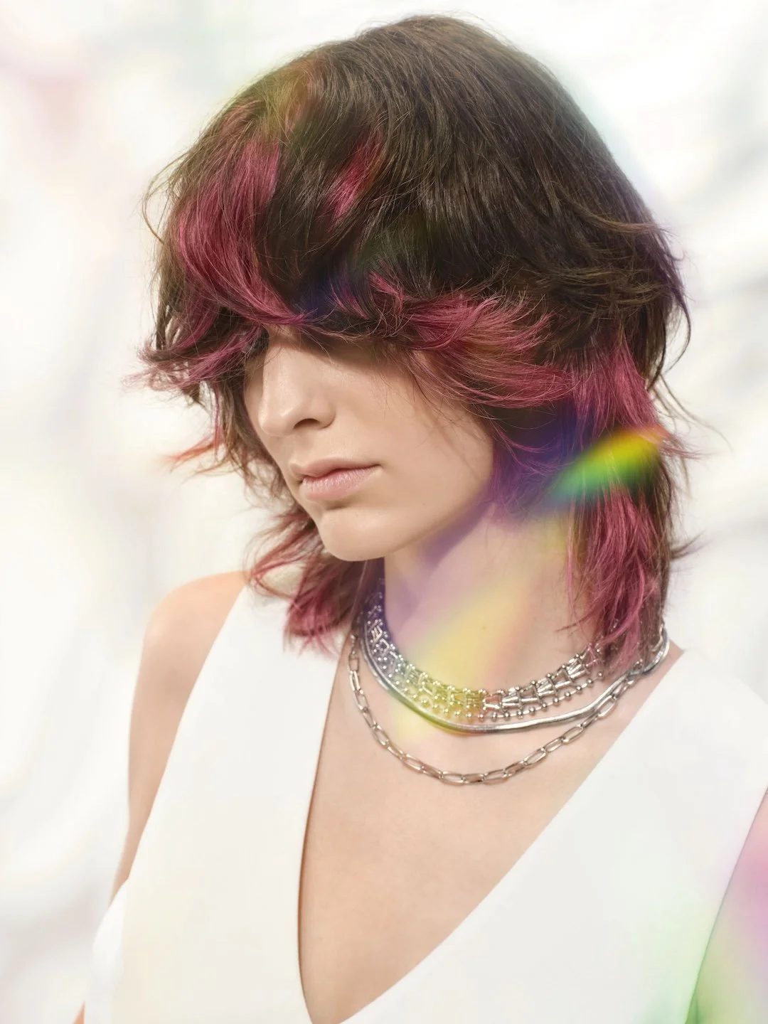 A woman with wavy, shoulder-length hair dyed pink and brown, wearing layered silver chain necklaces and a white sleeveless top, with a soft, blurry pastel background and rainbow-colored light flares.
