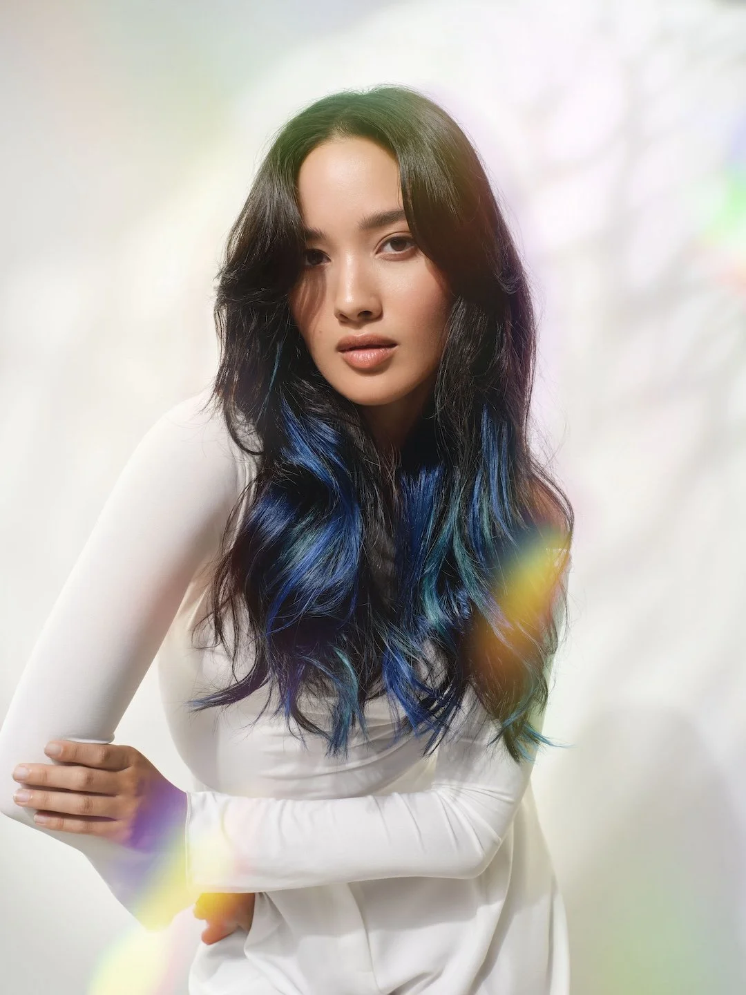 A woman with long, wavy black hair streaked with blue and green and wearing a white long-sleeve top poses with her arm crossed in front of her.