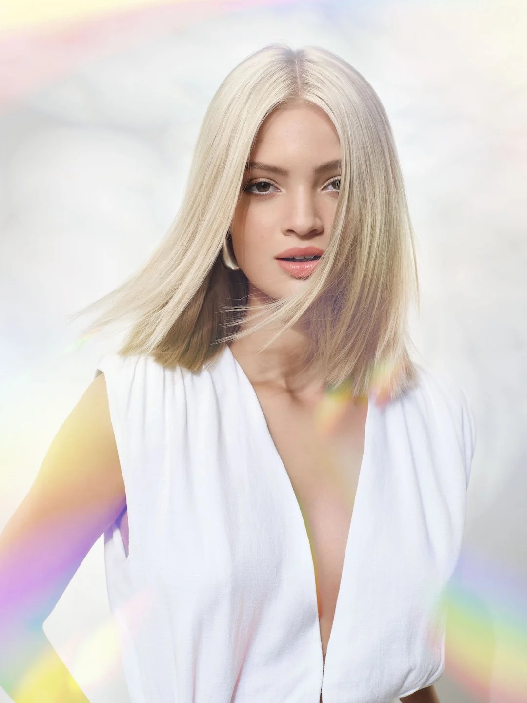 A young woman with blonde hair and light makeup wearing a white sleeveless top, looking into the camera against a soft, colorful background with rainbow light effects.