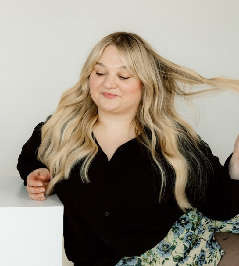 A woman with long blonde hair sitting and smiling with her eyes closed, wearing a black top and a floral skirt, against a plain light background.