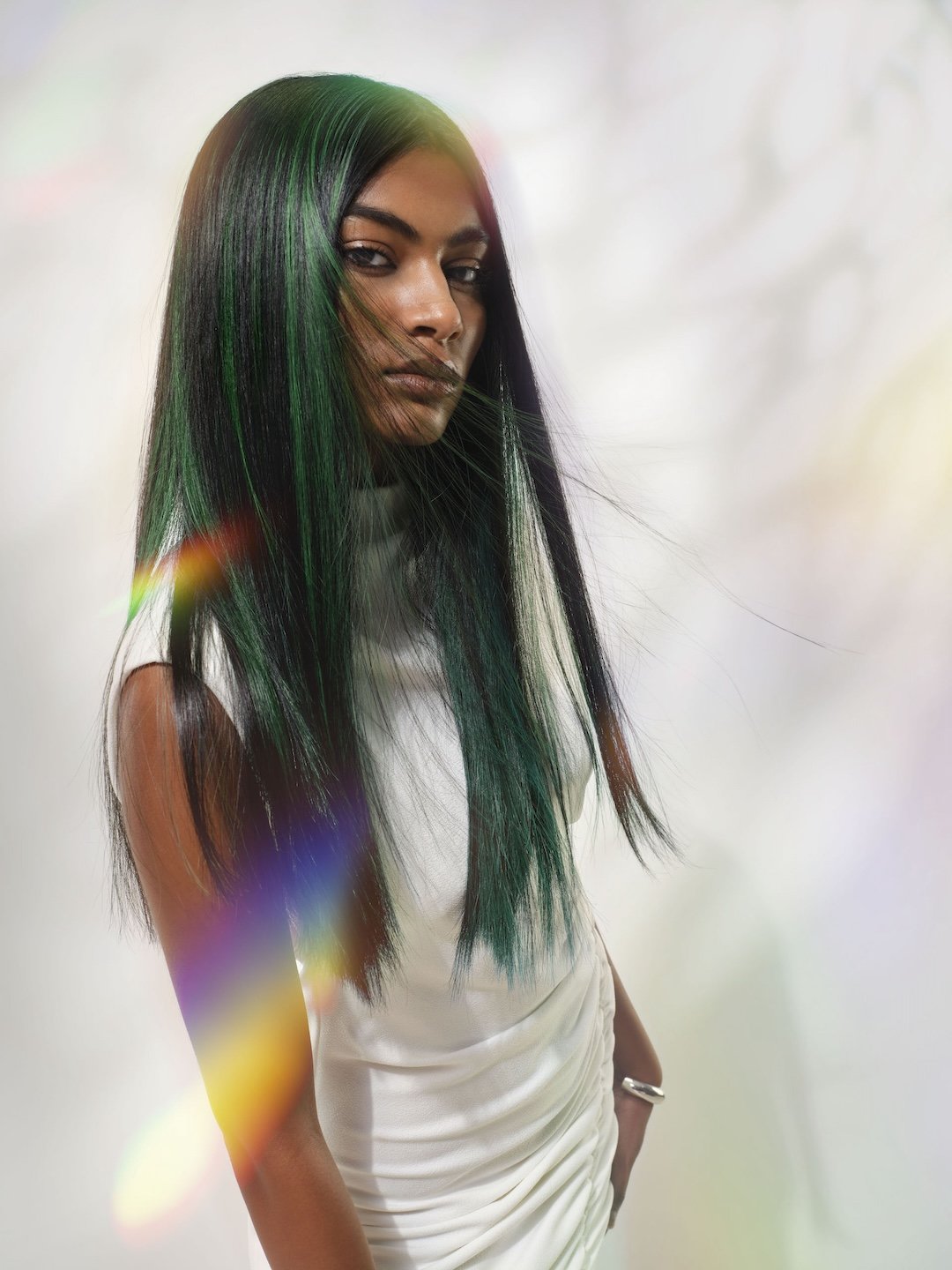 A woman with long, straight black hair with green highlights, wearing a white dress, standing against a plain white background with rainbow lens flare effects.