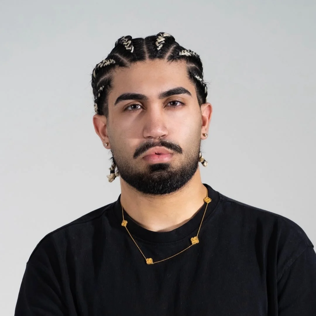 A young man with braided hair, a beard, and earrings wearing a black shirt and gold necklace against a plain background.