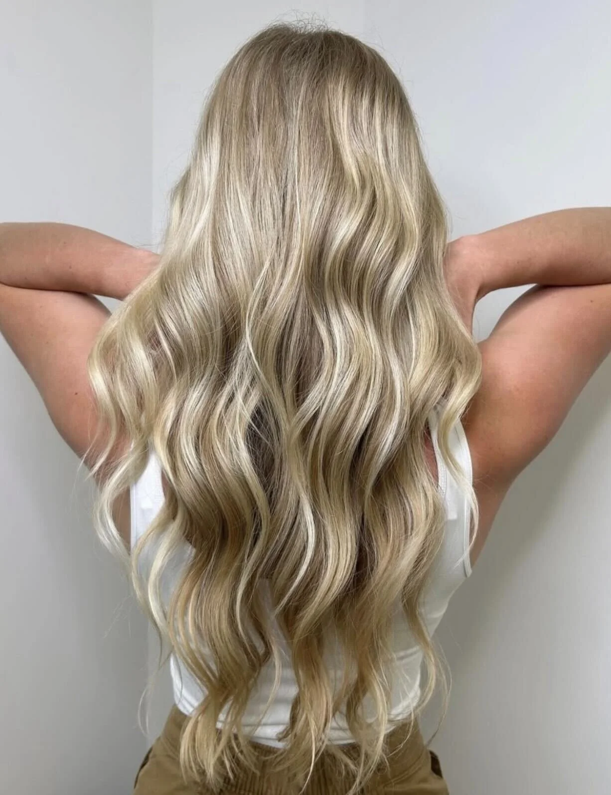Back view of a person with long, wavy blonde hair, standing against a light-colored wall, with arms raised and hands resting on the back of their head.