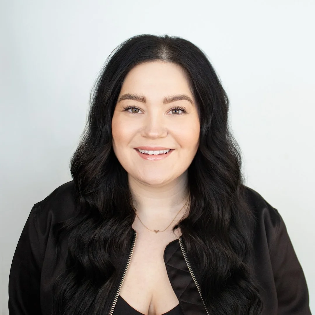 A woman with long black wavy hair, fair skin, smiling, wearing a black jacket and a delicate necklace, against a plain white background.