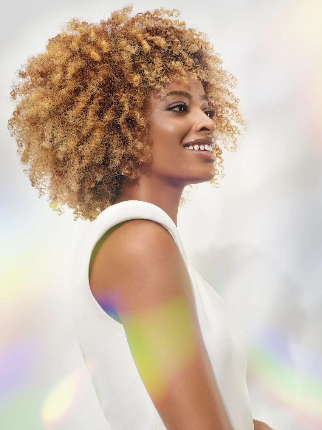 Side profile of a smiling woman with curly blonde hair wearing a white sleeveless top against a plain background.